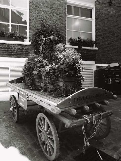 A wooden cart with large spoked wheels is parked on a brick-paved street outside a brick building with white-framed windows. The cart is loaded with a lush, dense arrangement of leafy plants and flowe