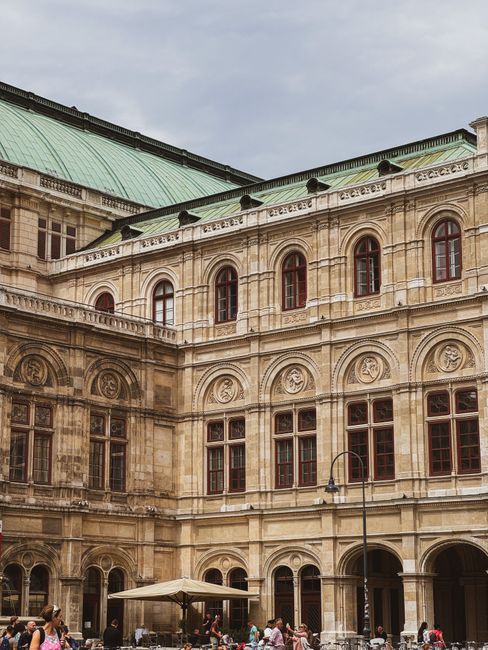 A historic building with ornate stonework and large arched windows, featuring a green, oxidized copper roof. The courtyard at the base of the building showcases landscaped garden beds with trimmed hed
