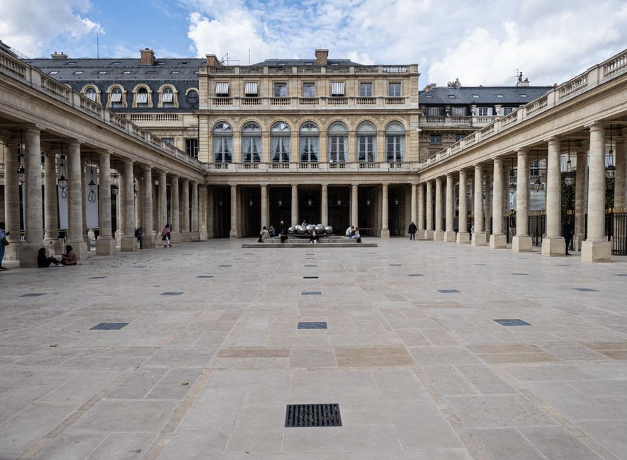 A historic building with ornate stonework and large arched windows, featuring a green, oxidized copper roof. The courtyard at the base of the building showcases landscaped garden beds with trimmed hed