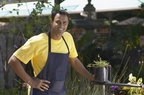 A smiling man wearing a yellow t-shirt and a dark apron stands in a well-maintained garden, holding a small black plant pot with fresh green foliage. The background features various garden plants, including ornamental grasses and shrubs, with a decorative stone fountain partially visible behind him. The garden area has a mix of paved and natural soil surfaces, surrounded by a wooden fence and bordered by flowering plants, shrubs, and greenery. The scene is lit by natural daylight, suggesting a bright, possibly partly cloudy day, emphasizing the lush, vibrant outdoor environment. This setting reflects professional gardening and outdoor maintenance services offered by Garden Maintenance Covent Garden in the local area.